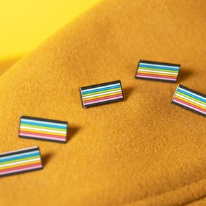 May include: Five rectangular enamel pins featuring a horizontal striped pattern of black, blue, light blue, white, yellow, pink, and red colors. Each pin has a thin black border surrounding the colorful stripes in a consistent design.