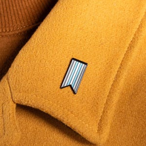 May include: A rectangular enamel pin with a swallowtail bottom edge, featuring vertical stripes in shades of light blue, white, and dark blue, attached to the collar of a mustard yellow wool coat worn over a brown ribbed turtleneck sweater.