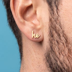 Peut inclure: Une boucle d'oreille à clou dorée en forme de lettres minuscules 'he' dans une police cursive. La boucle d'oreille est placée sur la partie inférieure du lobe de l'oreille, présentant une finition métallique lisse et polie au design simple.