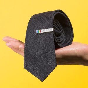 Puede incluir: Una corbata gris carbón oscuro con un acabado de tela texturizada, que presenta un pasador de corbata rectangular de tono plateado. El pasador está acentuado con una pequeña fila horizontal de cuadrados multicolores en rojo, amarillo, verde y azul.