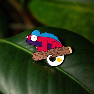 May include: A metal enamel pin shaped like a chameleon with a blue head, a red body, and a white tail featuring a small yellow heart. The chameleon is perched on a brown textured branch, depicted with a cartoon-like style and bold black outlines.