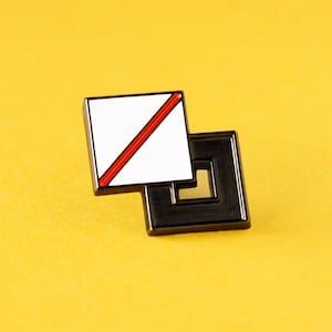 May include: A metal enamel pin featuring two overlapping square shapes. The top square is white with a single diagonal red stripe, while the bottom square is solid black with a hollow center, creating a geometric layered design.