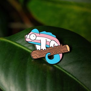 Könnte beinhalten: Eine Metall-Emaille-Anstecknadel in Form eines Chamäleons, das auf einem braunen Ast sitzt. Das Chamäleon zeigt horizontale Streifen in Hellblau, Rosa und Weiß, mit einem geringelten Schwanz und einem weißen Auge mit schwarzer Pupille.
