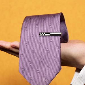 May include: A lavender necktie featuring a repeating diagonal pattern of small, subtle shapes. The tie is secured with a rectangular tie bar that displays a black and white checkered design on one side and a solid black finish on the other.