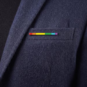 Könnte beinhalten: Eine rechteckige Anstecknadel mit einem horizontalen Regenbogenstreifenmuster in Rot, Orange, Gelb, Grün, Blau und Violett, befestigt an der Brusttasche eines strukturierten marineblauen Wollblazers, der über einem schwarzen Oberteil getragen wird.