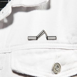 May include: A black and white enamel pin featuring a minimalist graphic of a bridge in an open position. The design consists of two horizontal segments with cross-hatch detailing and a central angled section representing a raised drawbridge.