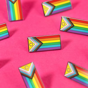 Könnte beinhalten: Eine Sammlung rechteckiger Emaille-Anstecknadeln mit dem Design der Progress Pride Flagge. Jede Anstecknadel zeigt horizontale Streifen in Rot, Orange, Gelb, Grün, Blau und Violett sowie ein Chevron-Muster in Weiß, Rosa, Hellblau, Braun und Schwarz auf der linken Seite.