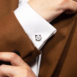 May include: A silver-toned metal cufflink featuring a circular face with a black arrow pointing upward and the text 01 A1 printed inside. The cufflink is secured to the white fabric cuff of a long-sleeved shirt worn under a brown wool blazer.