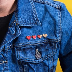 May include: A blue denim jacket featuring a row of four pixelated heart enamel pins attached above the chest pocket. The hearts transition from solid red to half-filled and empty, mimicking a classic video game health bar display.