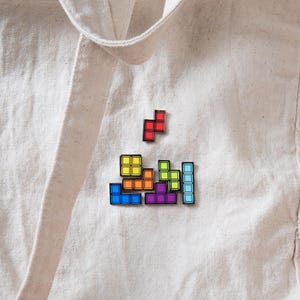 May include: A set of enamel pins shaped like colorful geometric game blocks attached to a beige canvas tote bag. The pins feature red, yellow, orange, green, purple, and light blue square segments arranged in various interlocking puzzle configurations.