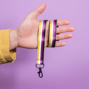 May include: A fabric lanyard featuring a striped pattern of yellow, white, purple, and black. The strap is attached to a black metal lobster clasp at the bottom, designed for holding items securely with a durable and functional metal hardware attachment.