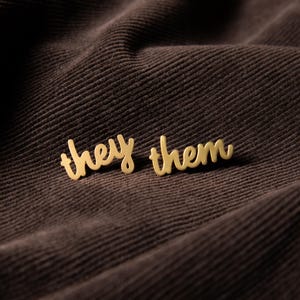 May include: A pair of gold-toned metal stud earrings featuring the words they and them written in a cursive script font. The earrings are separate pieces, with one displaying the word they and the other displaying the word them in lowercase.