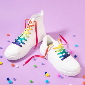 Könnte beinhalten: Ein Paar weiße Sneaker, ein High-Top und ein Low-Top, mit leuchtend regenbogenfarbenen Satinband-Schnürsenkeln. Die Schuhe sind von kleinen, bunten Pompons umgeben, die auf der Fläche verstreut sind und ein helles, verspieltes Bild ergeben.
