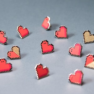 Puede incluir: Una colección de pequeños pines de esmalte con forma de corazón pixelado. Algunos pines presentan un color rojo brillante sólido, mientras que otros muestran un patrón de rayas diagonales en rojo y dorado. Cada pin tiene una estética digital.