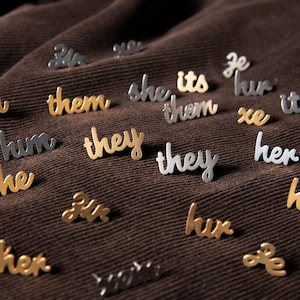 May include: A collection of small, individual metal pins shaped like pronouns in cursive script. The pins are finished in either a gold or silver tone and feature words such as they, them, she, he, her, him, its, and xe, scattered across the surface.