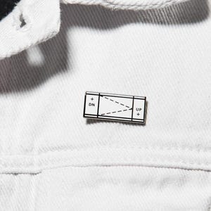 May include: A white denim jacket with silver metal buttons worn over a black shirt featuring a white grid pattern. A rectangular enamel pin is attached to the jacket, displaying a black and white diagram with the text SIN and COS.