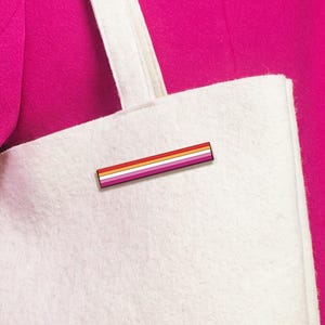 Könnte beinhalten: Eine weiße Filztasche mit einer rechteckigen Anstecknadel mit horizontalen Streifen in Orange-, Rosa- und Weißtönen. Die Tasche hat zwei Schulterriemen und eine strukturierte, weiche Oberfläche vor einem einfarbigen, leuchtend magentafarbenen Hintergrund.