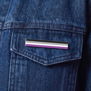 May include: A rectangular enamel pin featuring the asexual pride flag design with horizontal stripes in black, gray, white, and purple. The pin is attached to the chest pocket of a dark blue denim jacket worn over a dark blue button-down shirt.