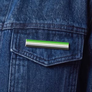 May include: A rectangular enamel pin featuring horizontal stripes in shades of green, white, gray, and black. The pin is attached to the chest pocket of a dark blue denim jacket, which is worn over a dark blue button-down shirt.