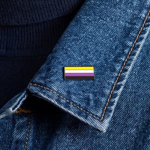 May include: A rectangular enamel pin featuring the non-binary pride flag with horizontal stripes in yellow, white, purple, and black. The pin is attached to the lapel of a blue denim jacket worn over a dark navy blue turtleneck sweater.
