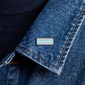 May include: A rectangular enamel pin featuring horizontal stripes in shades of light blue, white, and dark gray. The pin is attached to the collar of a blue denim jacket worn over a dark navy blue turtleneck sweater, showcasing a clean design.