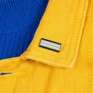May include: A rectangular enamel pin featuring horizontal stripes in black, white, gray, and light green. The pin is attached to the collar of a bright yellow denim jacket worn over a vibrant blue ribbed sweater against a solid cyan background.
