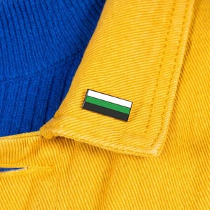 May include: A rectangular enamel pin featuring a horizontal tricolor design with a white top stripe, a green middle stripe, and a black bottom stripe. The pin is attached to the collar of a mustard yellow jacket worn over a blue ribbed shirt.