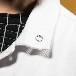 May include: A small circular enamel pin featuring a black geometric mountain-like design on a white background, attached to the collar of a white denim jacket worn over a black shirt with a white grid pattern.
