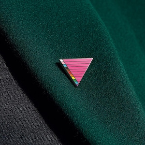 Pode incluir: Um broche triangular de esmalte com um campo rosa, linhas horizontais pretas e uma faixa vertical com gradiente de cores na borda esquerda, incluindo segmentos em branco, azul claro, castanho, preto, amarelo, verde e azul, fixado num tecido verde escuro.