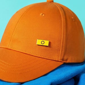 Può includere: Un cappellino da baseball arancione a tinta unita con una piccola spilla rettangolare gialla sul pannello frontale. La spilla presenta un'icona circolare nera al centro. Il cappellino ha una visiera curva e una corona strutturata con bottone.