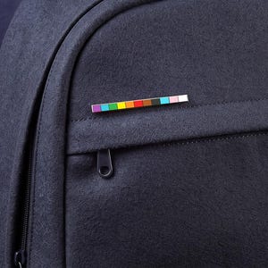 Könnte beinhalten: Ein dunkelblauer Rucksack aus Filz mit einer rechteckigen Emaille-Anstecknadel an der Vordertasche. Die Anstecknadel zeigt eine horizontale Reihe kleiner, leuchtender Quadrate in verschiedenen Farben, darunter Violett, Blau, Grün, Gelb, Orange, Rot, Braun, Schwarz, Hellblau und Pink.