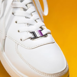 Op de afbeelding: Een witte sneaker met een rechthoekige metalen veterclip die aan de witte veters is bevestigd. De clip heeft een horizontaal streeppatroon in de kleuren zwart, grijs, wit en paars en is centraal geplaatst over de tong van de schoen.