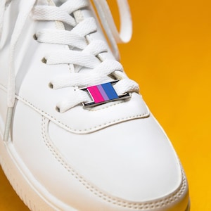 Pode incluir: Um tênis branco com um adorno metálico retangular para cadarços inserido nos cadarços brancos. O adorno apresenta um padrão de listras verticais nas cores rosa, roxo e azul, posicionado centralmente no sistema de amarração do calçado.
