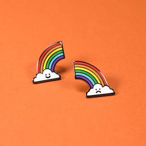 May include: Two metal enamel pins shaped like rainbows emerging from clouds. One pin features a white cloud with a smiling face, while the other pin features a white cloud with a sad face. Both rainbows display red, orange, yellow, green, blue, and purple stripes.