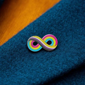 Puede incluir: Un broche de metal con forma de símbolo de infinito, que presenta rayas concéntricas de color rosa, amarillo y azul cian, con una banda central de color gris, blanco y negro. El broche está sujeto a una superficie de tela azul oscuro texturizada.