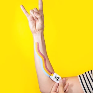 Puede incluir: Un aplicador de maquillaje rectangular blanco con el texto THE PIN PRICK impreso en el lateral. El aplicador est&aacute; trazando tres franjas onduladas paralelas de color azul claro, amarillo y rosa oscuro sobre un antebrazo con fondo amarillo.