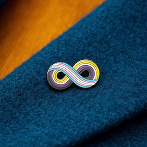 Può includere: Una spilla in smalto metallico a forma di simbolo dell'infinito. Il design presenta strisce concentriche gialle, bianche, viola, nere, azzurre e rosa, creando un effetto a strati. La spilla è fissata su un tessuto blu scuro testurizzato.