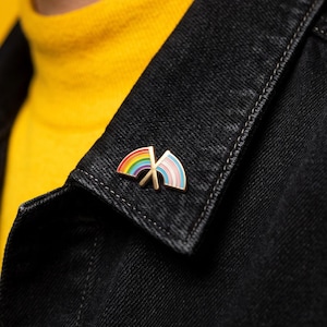 May include: A metal enamel pin featuring two crossed flag shapes. The left flag displays a horizontal rainbow stripe pattern, while the right flag features horizontal stripes in light blue, pink, and white. The pin is attached to a black denim collar.