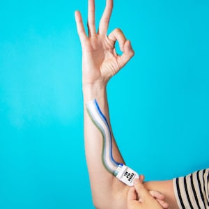 Puede incluir: Un aplicador de pl&aacute;stico blanco con la etiqueta THE PIN PRICK y una tira adhesiva a rayas azules, blancas y verdes que se extiende por el antebrazo. El dispositivo cuenta con un peque&ntilde;o mecanismo de rodillo en la base para aplicar la tira.