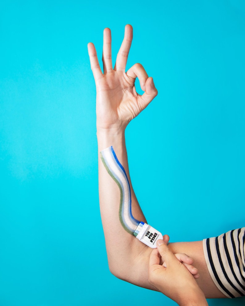 Puede incluir: Un aplicador de pl&aacute;stico blanco con la etiqueta THE PIN PRICK y una tira adhesiva a rayas azules, blancas y verdes que se extiende por el antebrazo. El dispositivo cuenta con un peque&ntilde;o mecanismo de rodillo en la base para aplicar la tira.