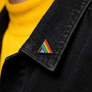 May include: A triangular enamel pin featuring a rainbow-coloured striped pattern with black, brown, white, pink, and light blue accents. The pin is attached to the collar of a black denim jacket worn over a bright yellow shirt.