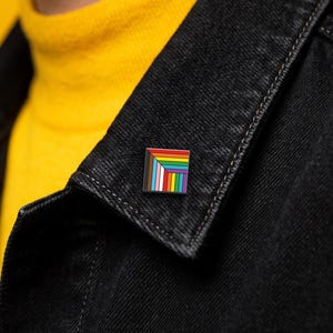 May include: A square enamel pin featuring a geometric design with a series of nested right-angled stripes in black, brown, light blue, white, pink, red, orange, yellow, green, and purple, arranged in a corner-aligned pattern on a dark metal base.