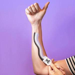 Puede incluir: Un aplicador manual blanco con la etiqueta THE PIN PRICK que aplica una franja ondulada de tres tonos de pigmento negro, gris y morado claro sobre la piel. El dispositivo tiene un cuerpo rectangular con una punta texturizada para crear una l&iacute;nea de color continua.