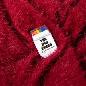 Puede incluir: Un recipiente rectangular de pl&aacute;stico blanco con una tapa transparente que revela cinco peque&ntilde;os l&aacute;pices de colores en rojo, naranja, amarillo, verde y azul. El frente presenta el texto negro THE PIN PRICK y el usuario de redes sociales @PINPRICK.SHOP.