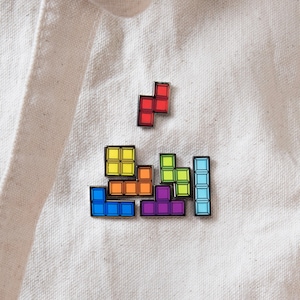 May include: A collection of enamel pins shaped like colorful geometric block puzzle pieces. The set includes a red piece, a yellow square, an orange L-shape, a blue L-shape, a purple T-shape, a lime green piece, and a light blue vertical bar.