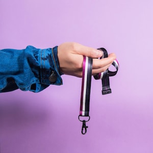 May include: A fabric lanyard featuring a vertical striped pattern in magenta, white, and black. The lanyard includes a black metal lobster clasp at the bottom end and a black plastic connector piece, designed for holding keys or identification badges.