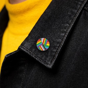 May include: A circular enamel pin featuring a vibrant rainbow-coloured pattern with intersecting diagonal lines. The design incorporates multiple bright hues including red, orange, yellow, green, blue, and purple, arranged in a geometric, multi-directional striped motif.