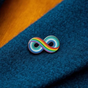 May include: A metal enamel pin shaped like an infinity symbol, featuring concentric rings of rainbow colours including red, orange, yellow, green, light blue, and dark blue, with a thin silver-toned border outlining the entire shape.