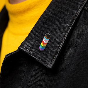 May include: A rectangular enamel pin featuring a vertical stack of coloured stripes in black, brown, light blue, pink, white, red, orange, yellow, green, and purple, attached to the collar of a black denim jacket worn over a yellow shirt.