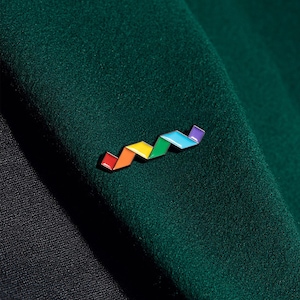 Puede incluir: Un broche de esmalte en forma de zigzag que presenta una secuencia de cinco segmentos de colores en rojo, naranja, amarillo, verde y azul, delineados en metal negro. El broche está sujeto a una superficie de tela verde oscuro con textura.